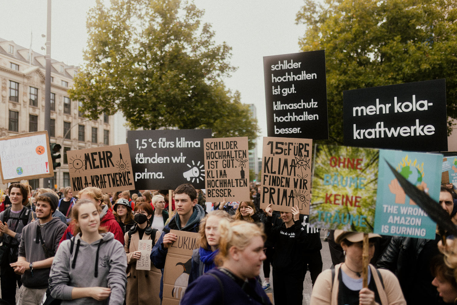 menschen demonstrieren auf einer straße und halten verschiedene schilder mit schriftzügen und illustrationen, bäume und gebäude im hintergrund