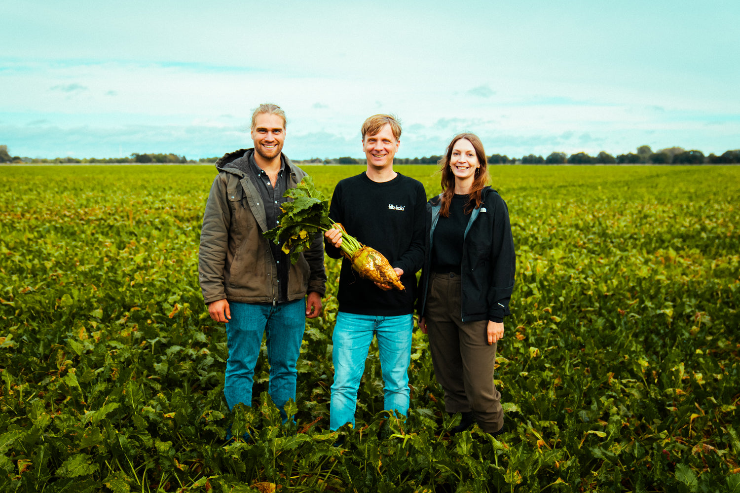 drei personen stehen auf einem feld mit grünen pflanzen, eine person in der mitte hält eine große pflanze mit wurzel in die kamera, freies feld und himmel im hintergrund