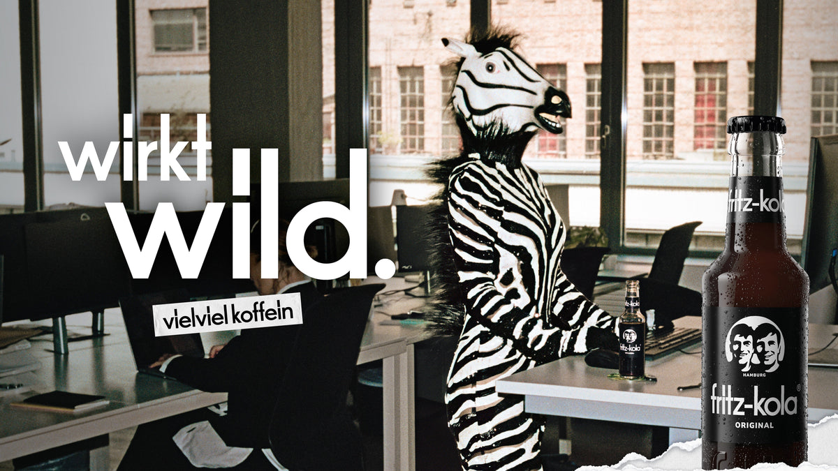 fritz-kola Kampagnenmotiv „wirkt wild.“ mit Person im Zebrakostüm im Büro und fritz-kola original Flasche.