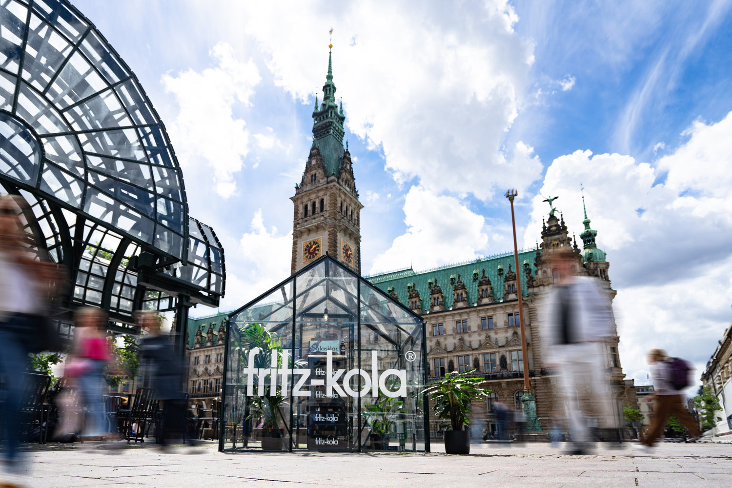 gläserner pavillon mit fritz-kola schriftzug auf einem platz vor einem historischen gebäude mit turm, menschen bewegen sich unscharf im vordergrund