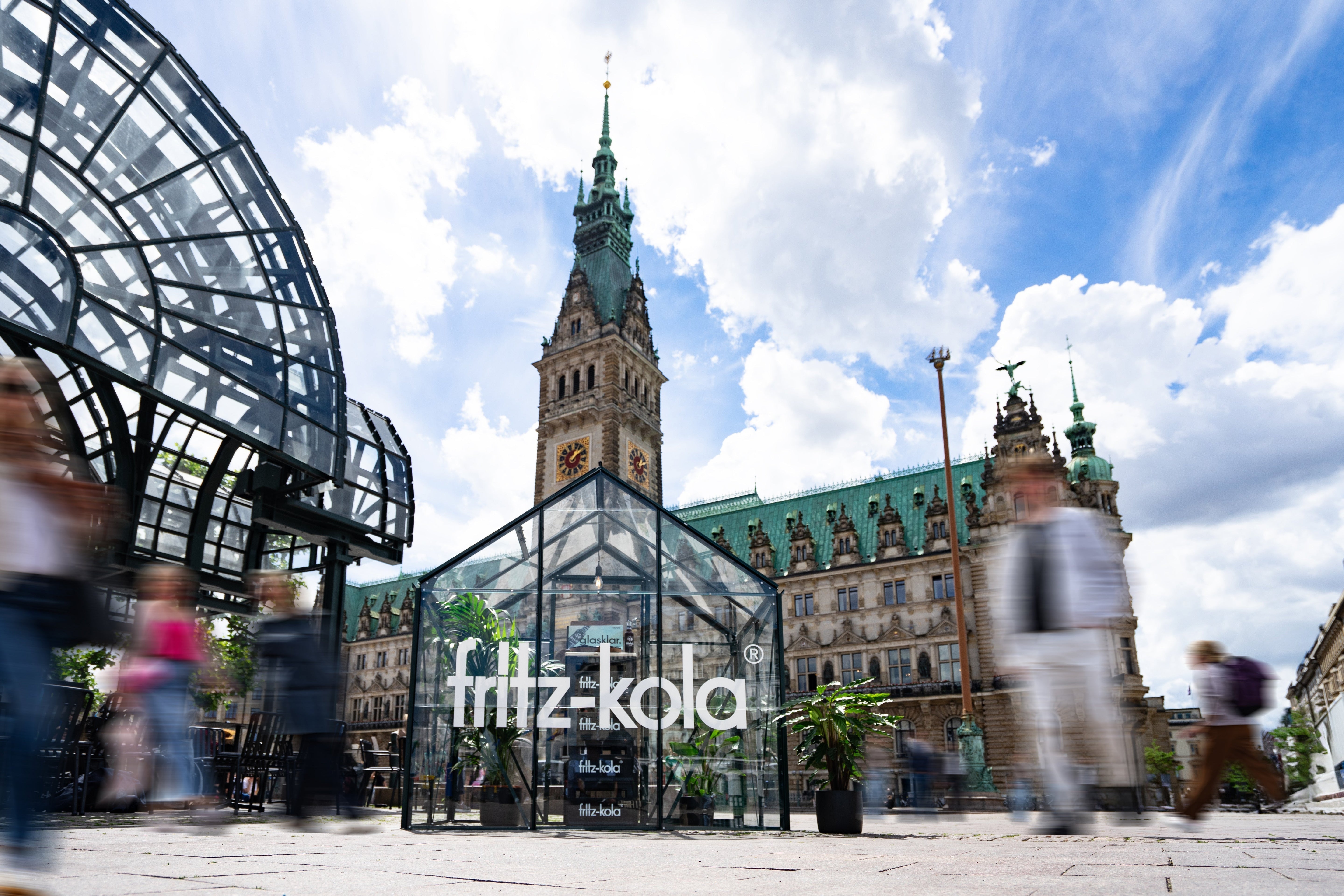 gläserner pavillon mit fritz-kola schriftzug auf einem platz vor einem historischen gebäude mit turm, menschen bewegen sich unscharf im vordergrund