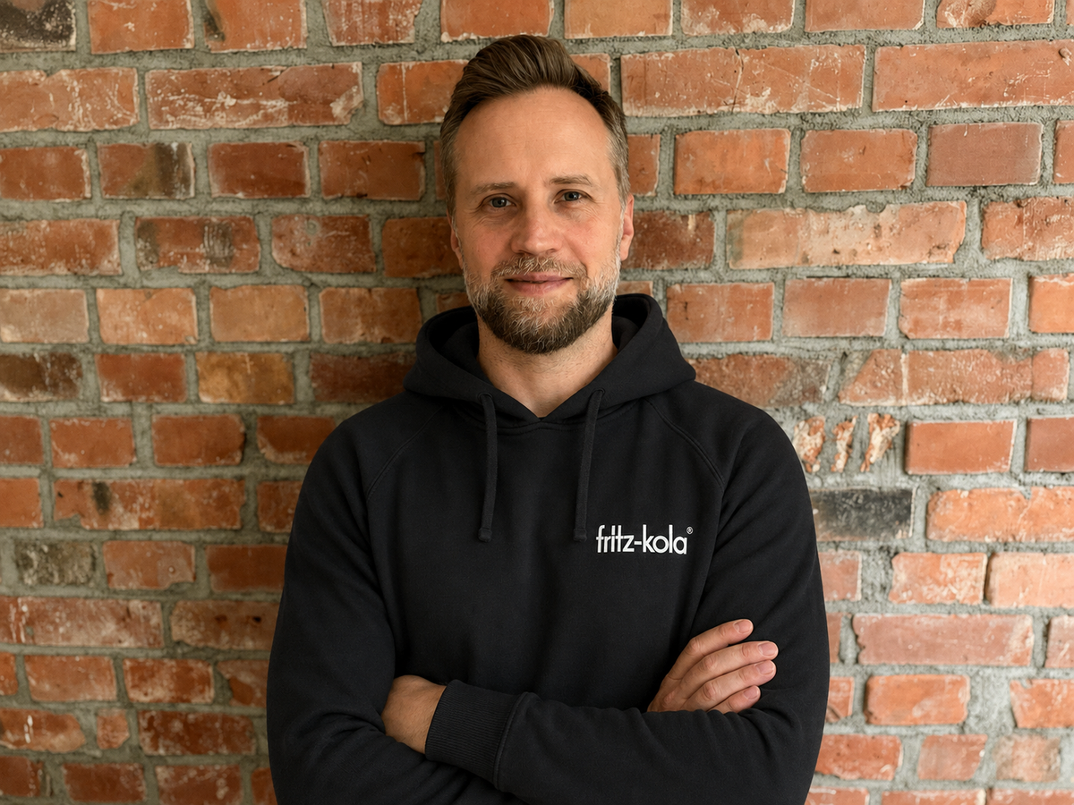 Marcin Piaseczny, new marketing director of fritz-kola, in a black fritz-kola Hoodie in front of a brick wall.