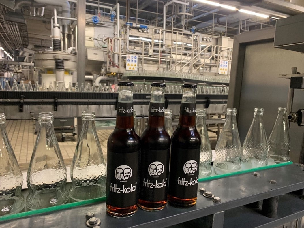 drei fritz-kola glasflaschen stehen auf einem förderband zwischen leeren glasflaschen in einer industriellen abfüllanlage, maschinen und rohre im hintergrund