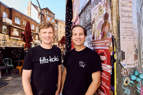 Vor einer plakatierten Wand in einer städtischen Umgebung stehen die fritz-kola Geschäftsführer Mirco Wolf Wiegert und Florian Weins lächeln nebeneinander.