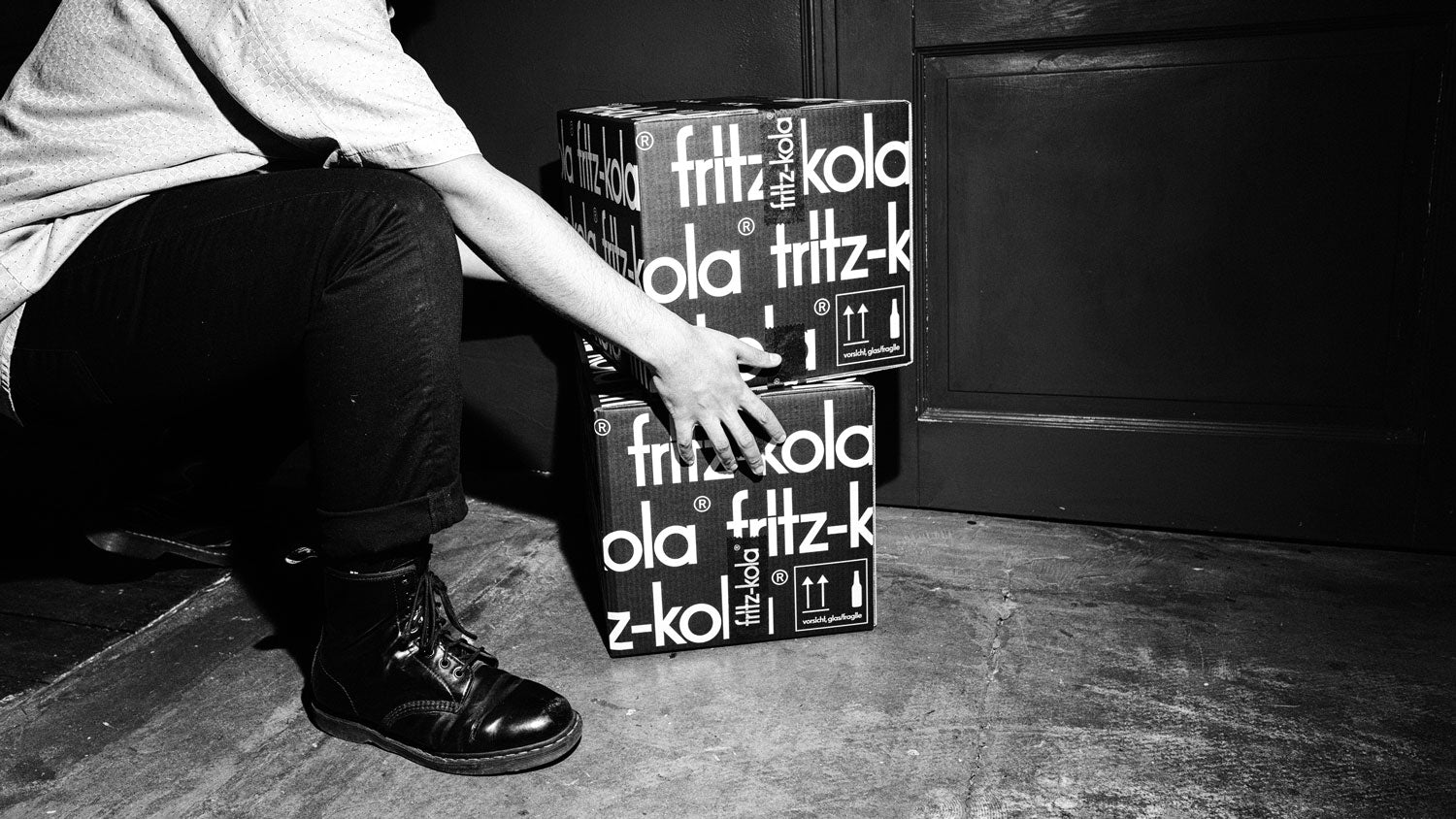 Schwarz-weiß Foto: Person in hellem Hemd und schwarzen Schuhen stapelt zwei schwarze fritz-kola Kartons mit weißem Schriftzug.