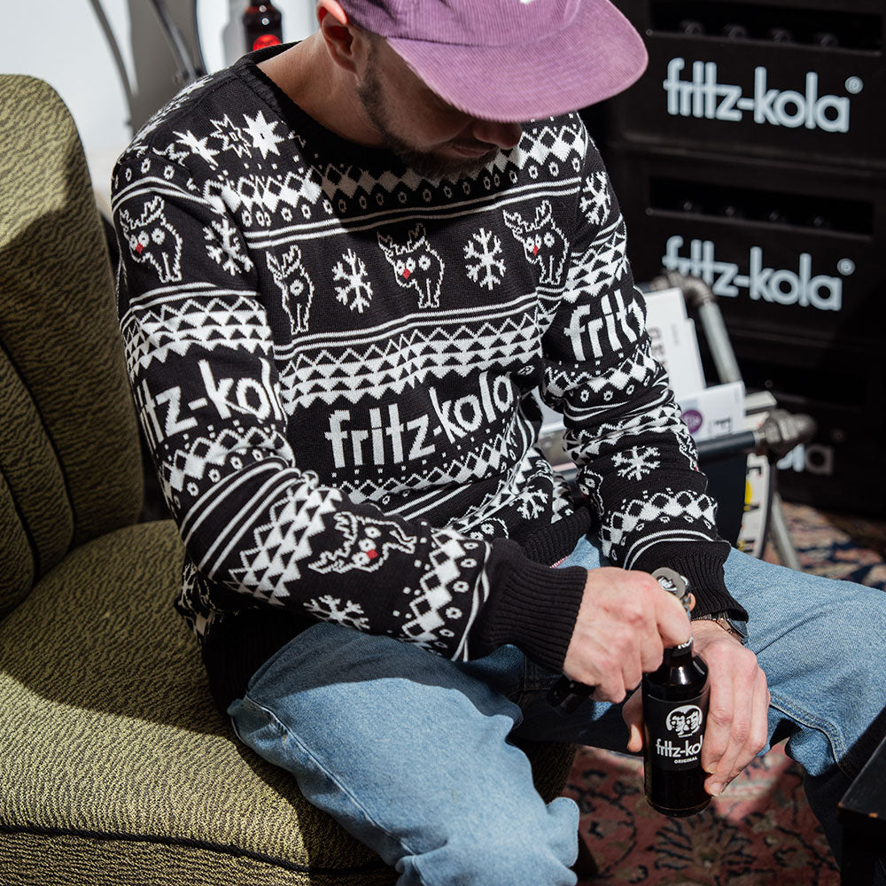 Eine Person sitzt auf einem Sessel und trägt einen weihnachtlichen fritz-kola Strickpullover im skandinavischen Design mit Rentieren und Schneeflocken in Schwarz-Weiß. Die Person trägt eine violette Kappe und hält eine fritz-kola Flasche in den Händen. Sie trägt eine helle Jeans und ist vor fritz-kola Getränkekisten fotografiert.