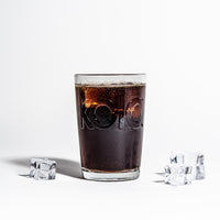 Ein mit fritz-kola gefülltes Glas, flankiert von Eiswürfeln auf beiden Seiten. Die dunkle Cola kontrastiert stark mit dem weißen Hintergrund. Der eingeprägte kola Schriftzug ist deutlich sichtbar, die Oberfläche des Glases zeigt Kondensation.
