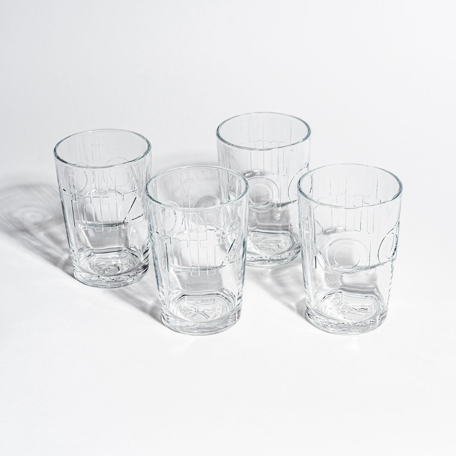 Vier leere fritz-kola Gläser auf weißem Hintergrund, fotografiert von oben. Die Gläser sind in quadratischer Anordnung platziert und zeigen deutlich die eingeprägten fritz-kola Schriftzüge. Klares Kristallglas mit charakteristischen Rillen und Prägungen wirft dezente Schatten.