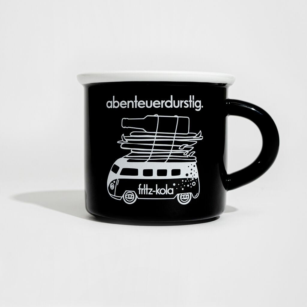 Schwarze Tasse mit weißem Text "abenteuerdurstig." oben und "bretterfritzen" unten. In der Mitte steht ein gezeichneter VW-Bus mit Surfbrettern auf dem Dach und einer fritz-kola Flasche obendrauf. Der Bus hat Fenster und Graffiti-artige Verzierungen an den Seiten mit Meereselementen wie Fischen und Wellen.
