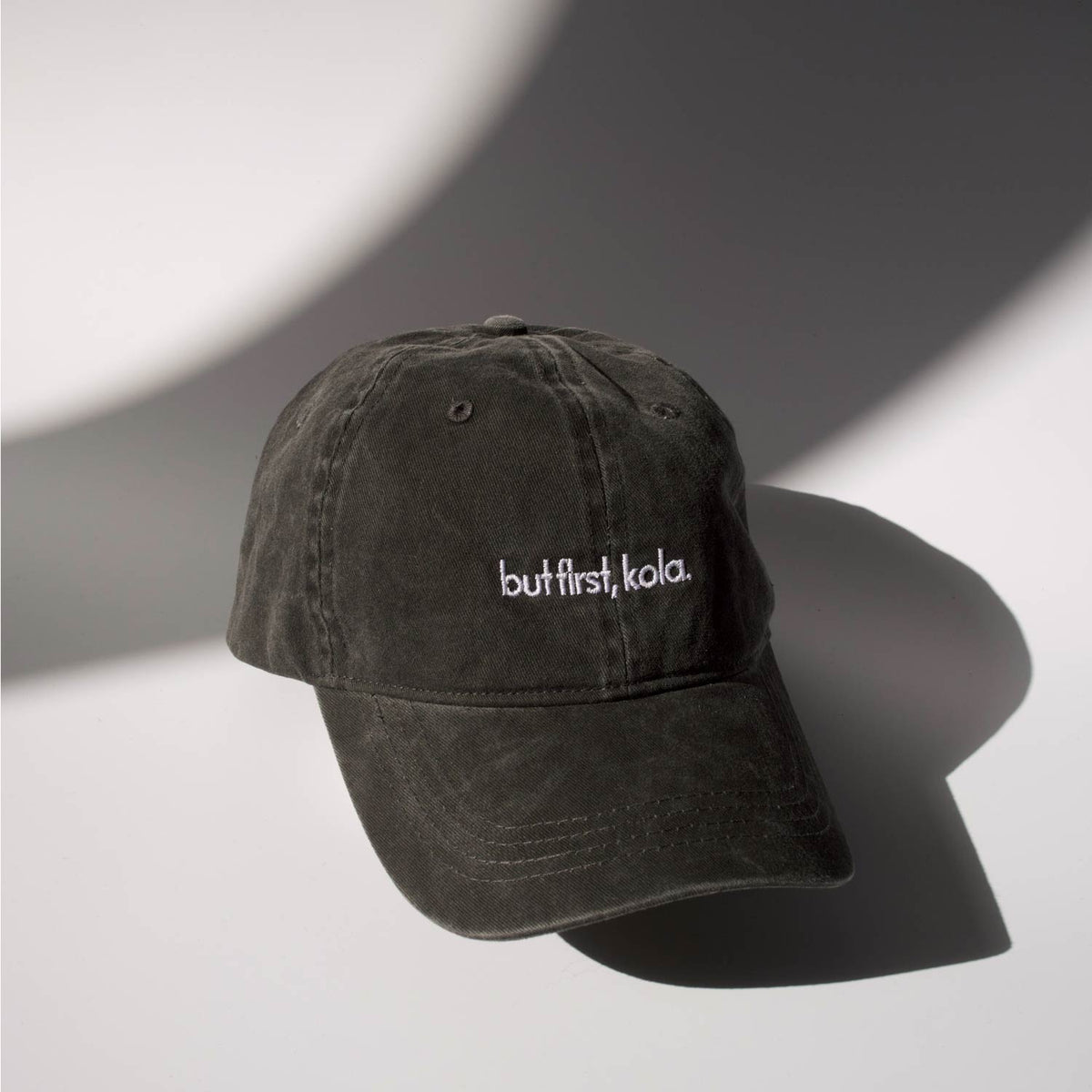 Schwarze Cap in verwaschener Optik mit weiß gesticktem Schriftzug „but first, kola.“ auf der Vorderseite, fotografiert vor hellem Hintergrund mit weichen Licht- und Schattenflächen.