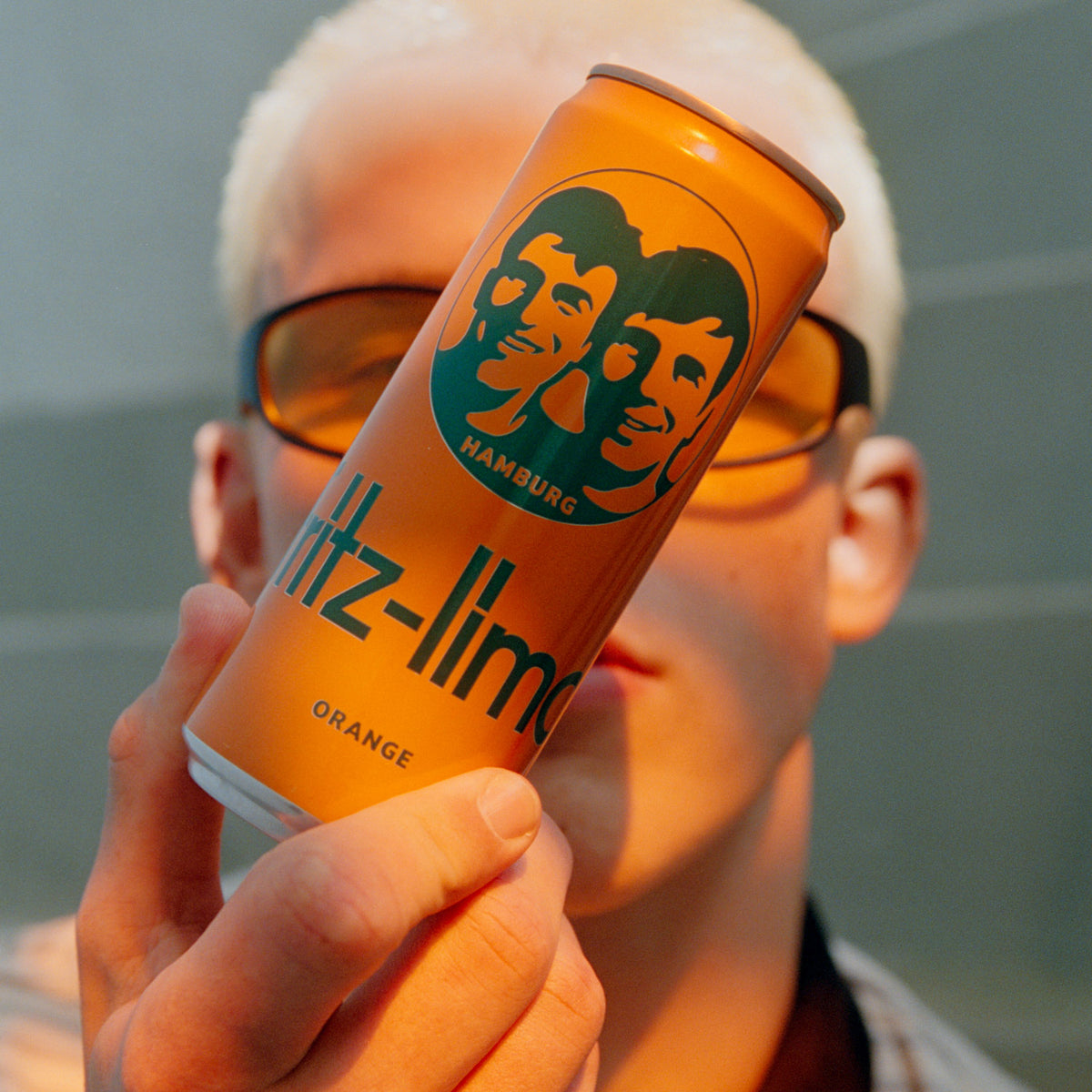 Person hält eine Dose fritz-limo Orange vor das Gesicht. Die orangefarbene Dose steht im Fokus, während die Person mit orange getönten Sonnenbrillen im Hintergrund unscharf zu sehen ist.