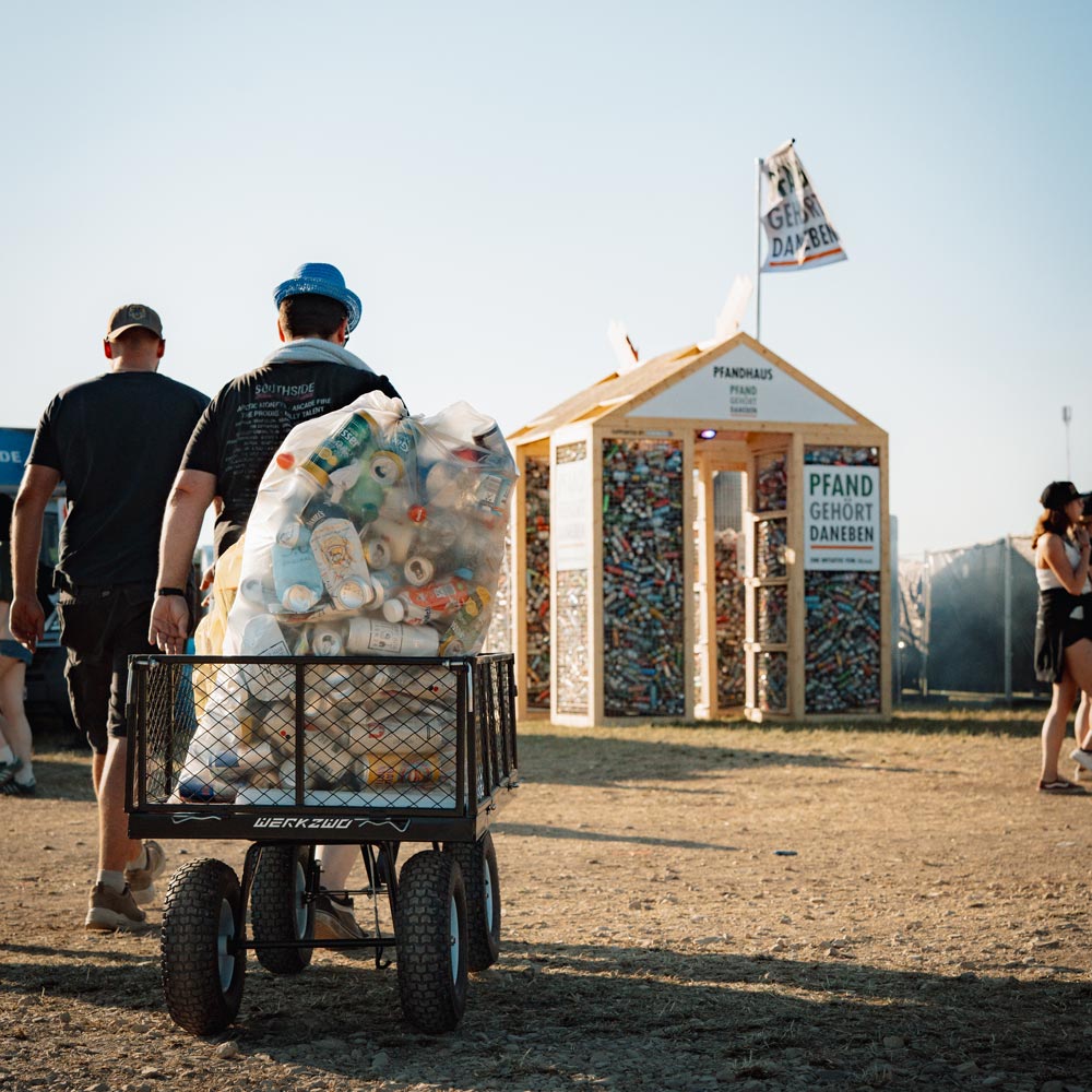 Zwei Festivalbesucher schieben einen Bollerwagen mit weißen Müllsäcken voller Leergut über staubigen Boden. Im Hintergrund steht das Pfandhaus - ein kleines Holzhaus mit Dachfahne "Pfand gehört daneben". Die Außenwände sind voller bunter Getränkedosen hinter Gittern.