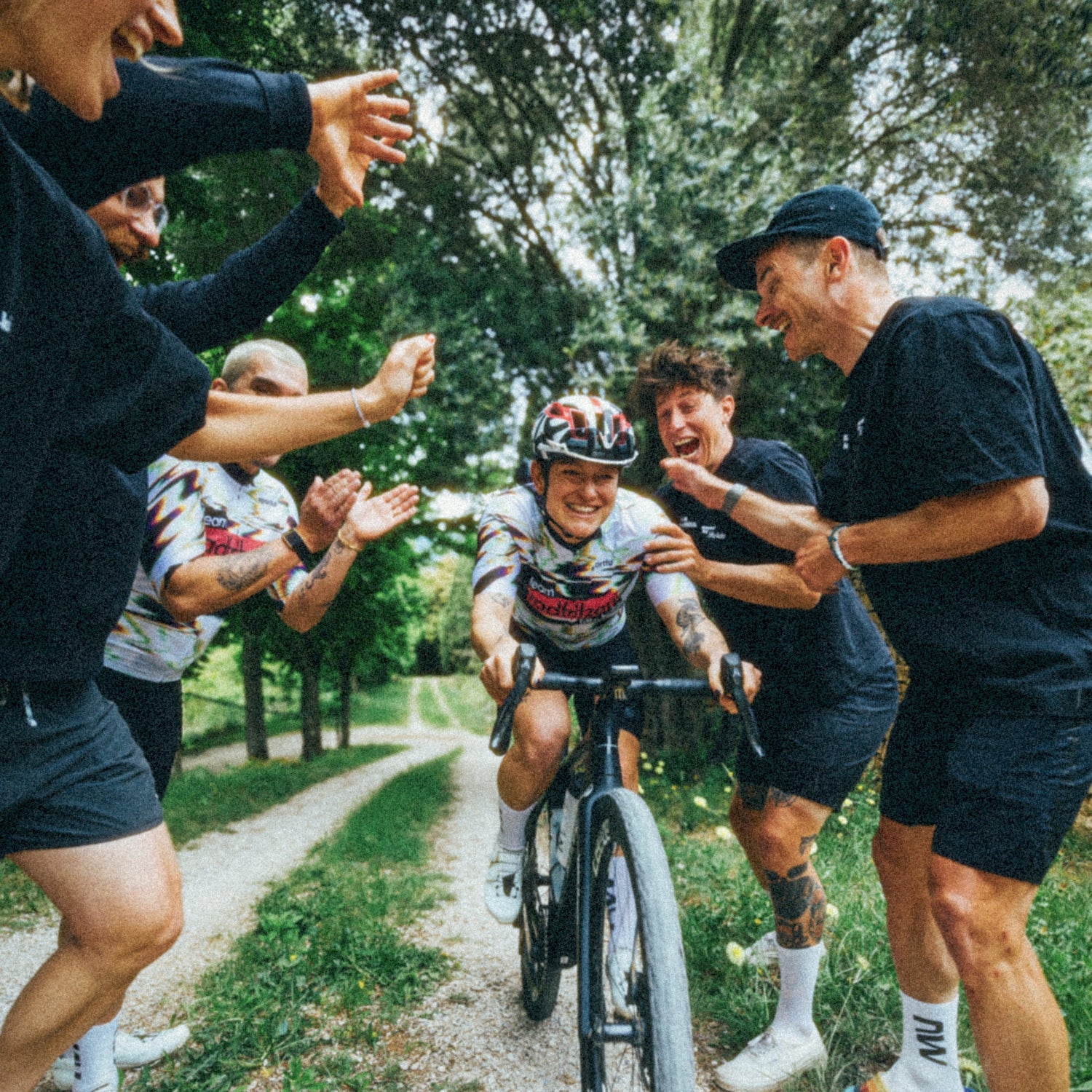 Person auf Rennrad trägt radfritzen Trikot und fährt auf einem schmalen Naturweg, umgeben von mehreren Personen, die applaudieren und anfeuern; grüner Park mit Bäumen im Hintergrund.