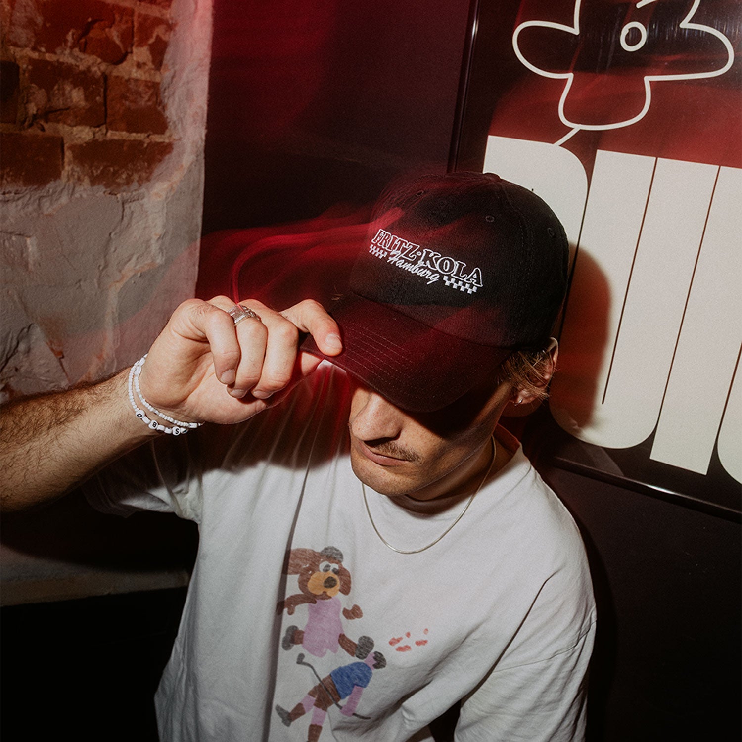 Mann in weißem Shirt trägt schwarze fritz-kola Cap. Fasst mit der Hand an den Schirm, warmes rotes Licht beleuchtet die Szene vor Backsteinwand.