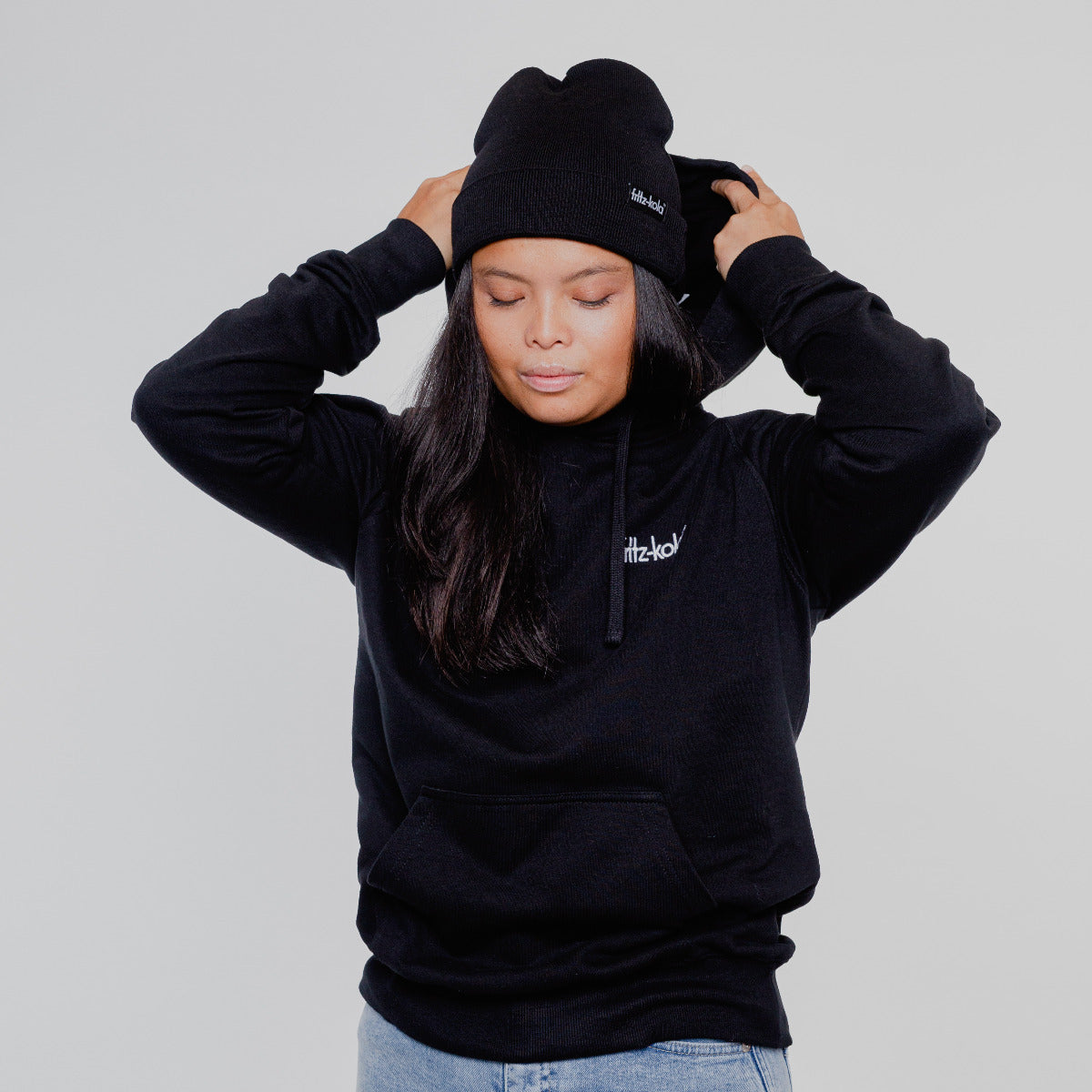 Frau in schwarzem Kapuzenpullover mit weißem fritz-kola Schriftzug auf der Brust. Sie trägt eine schwarze beanie ebenfalls mit fritz-kola schriftzug.