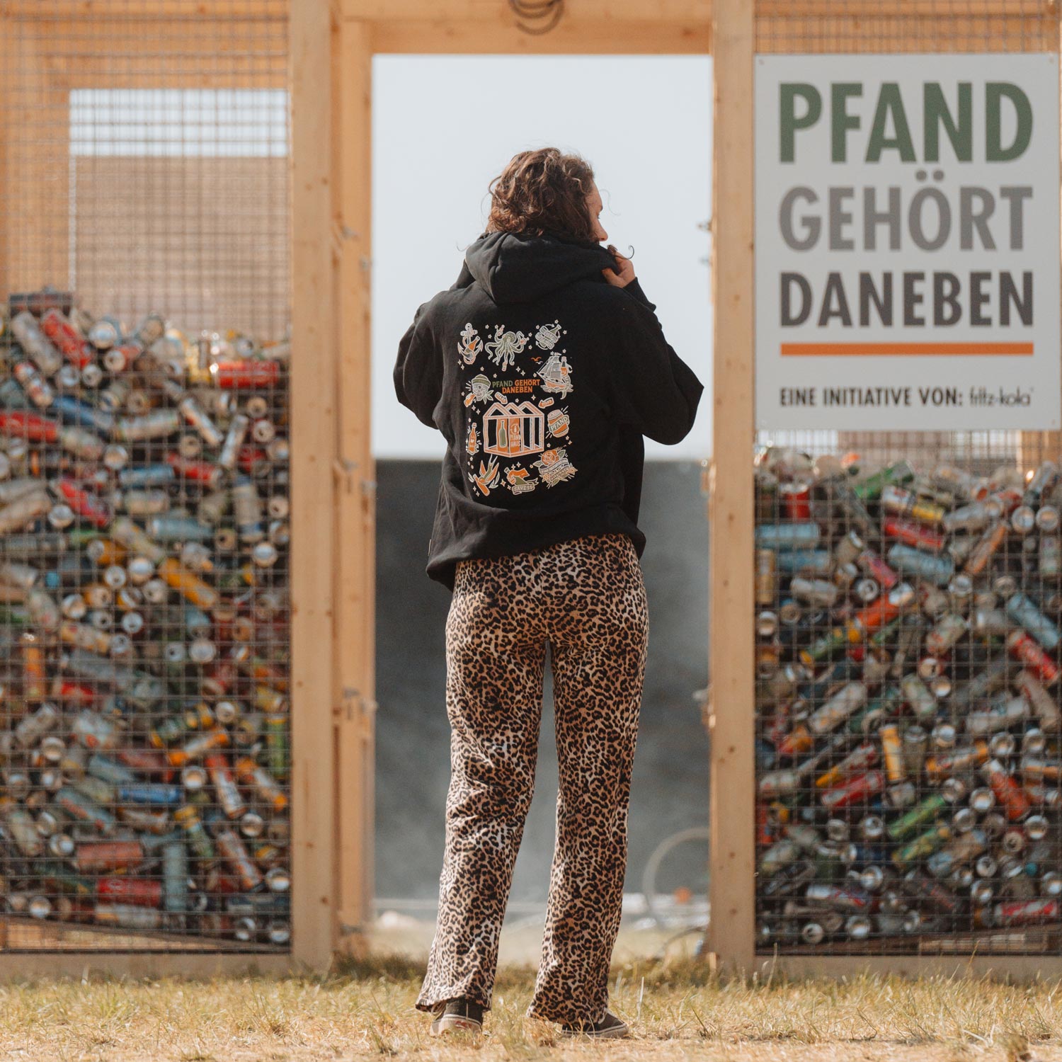 Eine Person in schwarzem Hoodie mit Leopardenmuster-Hose steht vor dem hölzernen Pfandhaus auf einem Festival. Das Pfandhaus zeigt das Schild "PFAND GEHÖRT DANEBEN" und ist mit gesammelten Pfandflaschen gefüllt. Auf dem Rücken des Hoodies ist das bunte Design der "Pfand gehört daneben"-Kollektion zu sehen, das maritime und Pfand-Motive vereint.