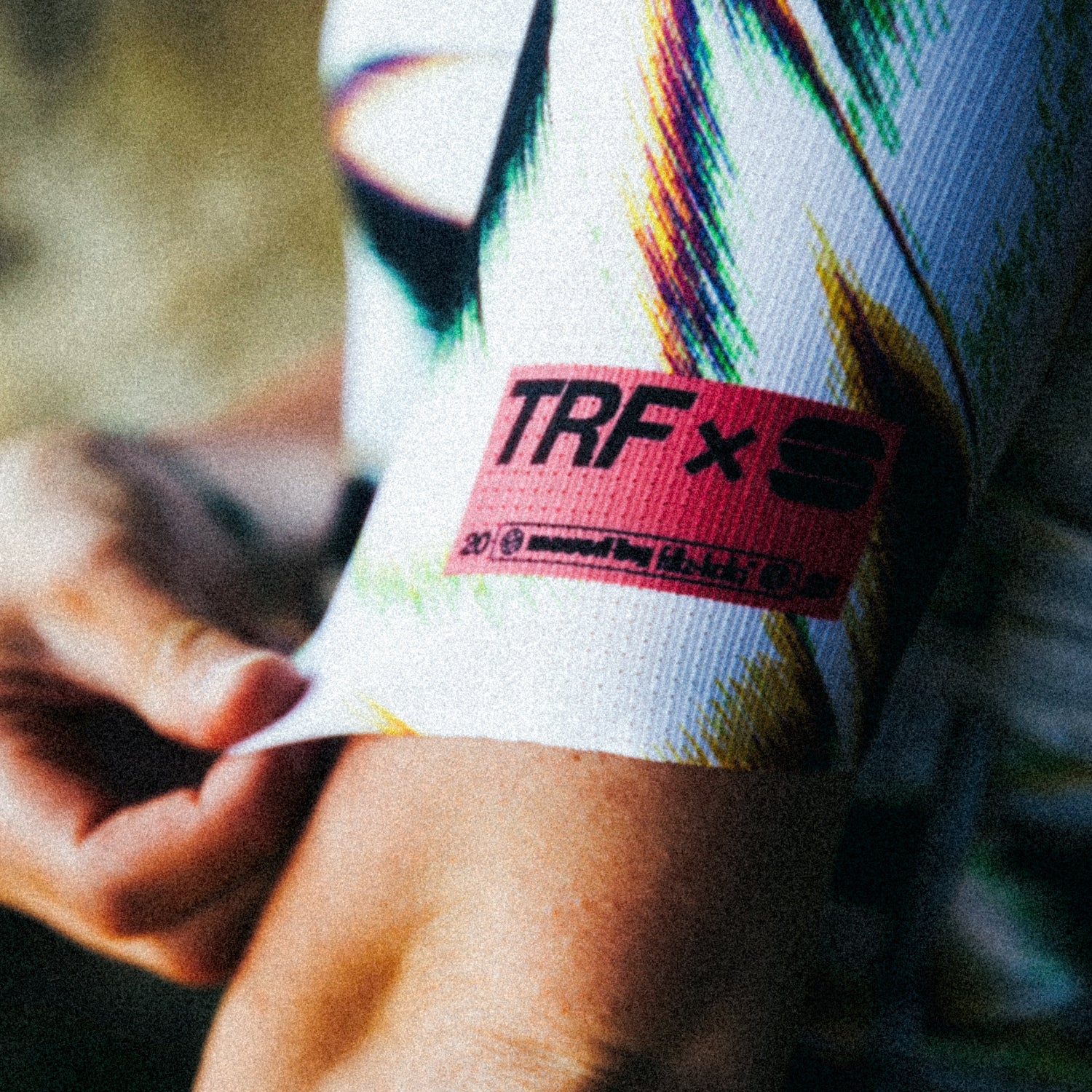 Nahaufnahme des radfritzen Trikots am Arm mit farbigem Muster und pinkem „TRF“ Patch; Person zieht den Ärmel leicht nach unten.