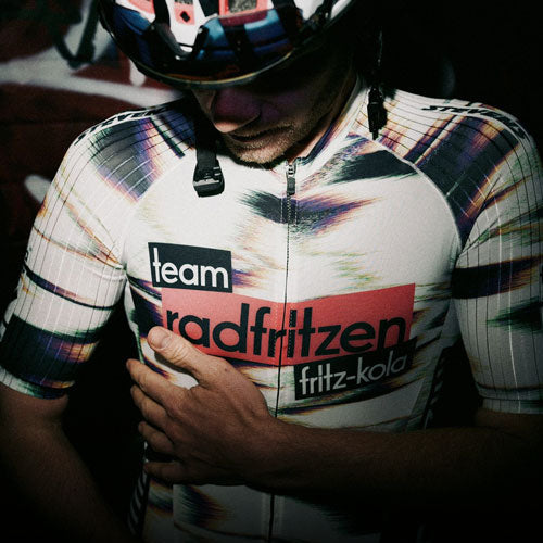 Nahaufnahme einer Person in Fahrradkleidung und Helm. Auf dem Trikot steht „team radfritzen“ sowie „fritz-kola“. Eine Hand liegt auf der Brust, der Hintergrund ist dunkel und unscharf.