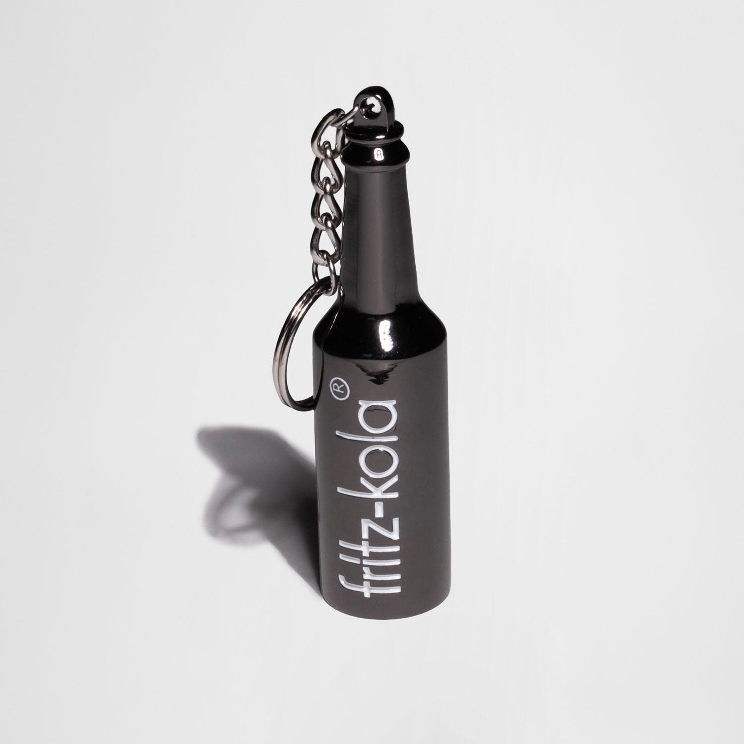 Schlüsselanhänger aus dem dunklem Metall in Form einer Flasche vor hellem Hintergrund mit Schlagschatten Fotografiert. Weißes fritz-kola logo auf der Flasche