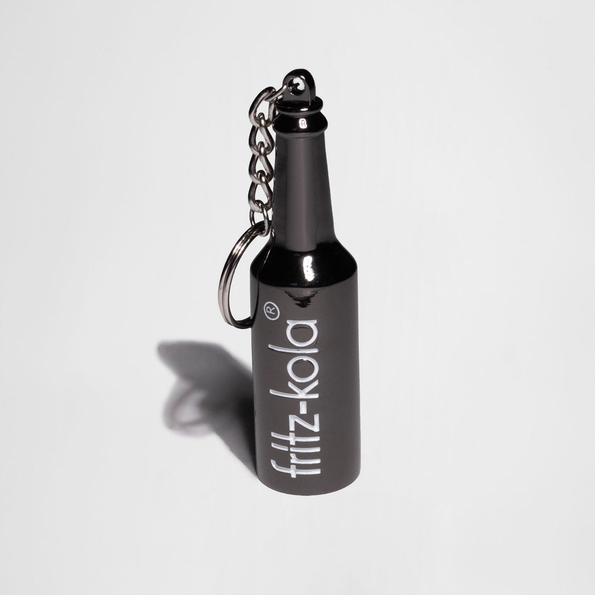 Schlüsselanhänger aus dem dunklem Metall in Form einer Flasche vor hellem Hintergrund mit Schlagschatten Fotografiert. Weißes fritz-kola logo auf der Flasche