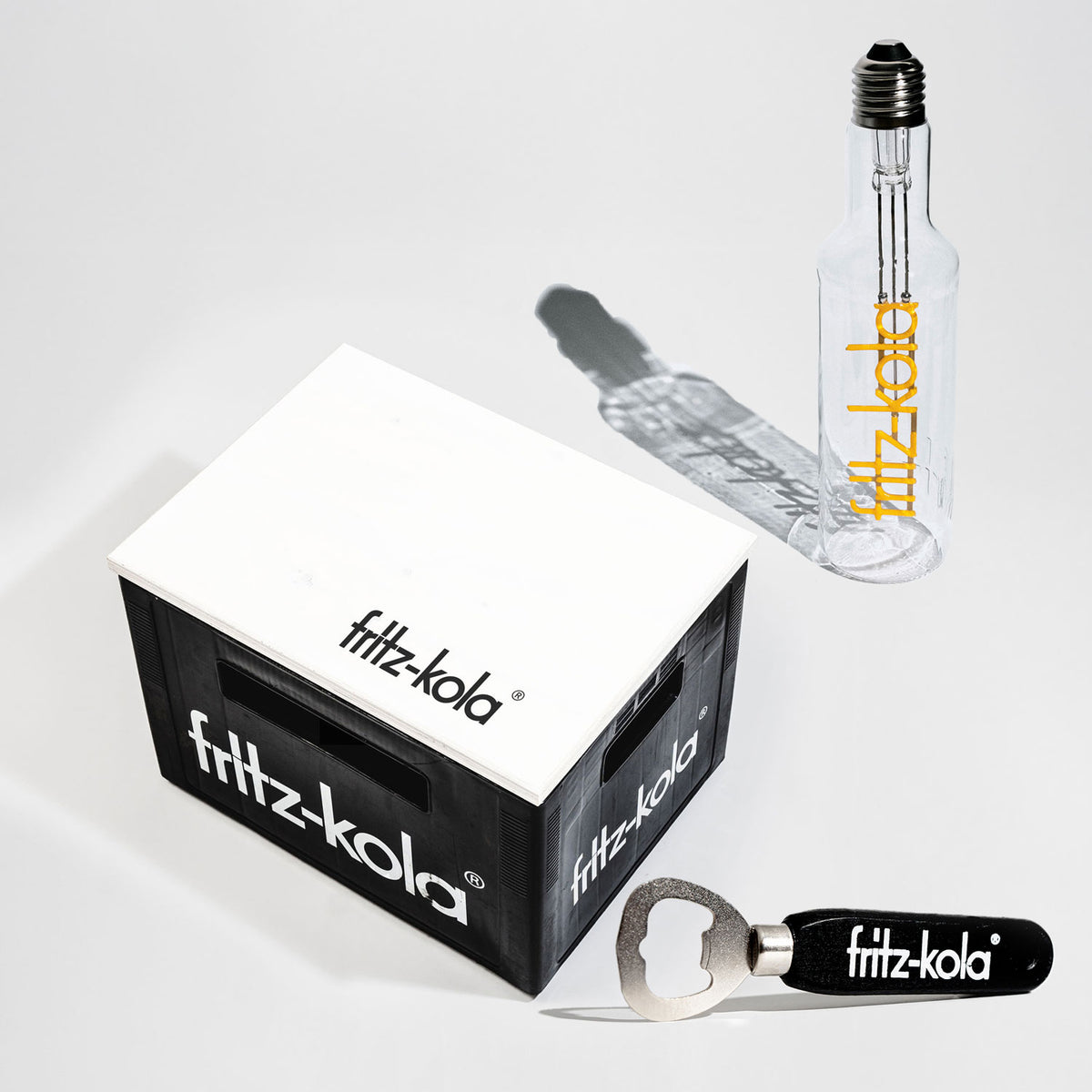 Set aus fritz-kola Merchandise für Zuhause: schwarze Mini-Kiste mit weißem fritz-kola-Logo, transparente Glasflasche im Glühbirnen-Design mit gelbem fritz-kola-Schriftzug und ein schwarzer Flaschenöffner mit weißem fritz-kola-Logo.