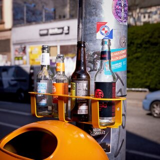 Ein an einer Laterne befestigter Mülleimer hat über dem Deckel ein Metallständer befestigt, welcher mehrere Pfandflaschen hält