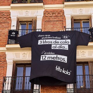 Una camiseta negra gigante con un eslogan publicitario de fritz-kola cuelga de un balcón en una fachada de ladrillo. El texto en la camiseta dice: “Claro que puedes beber 3 litros de cola al día… si mides 12 metros.
