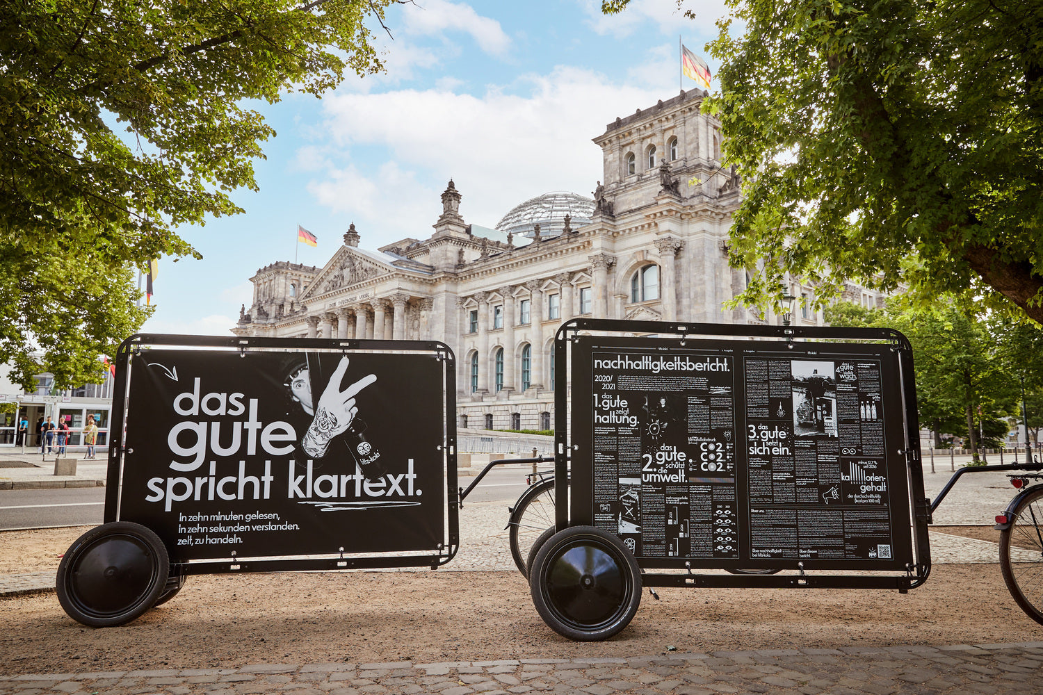 zwei anhänger mit großflächigen schwarz-weißen plakaten stehen auf einem platz vor einem historischen gebäude mit kuppel, bäume rahmen die szene ein