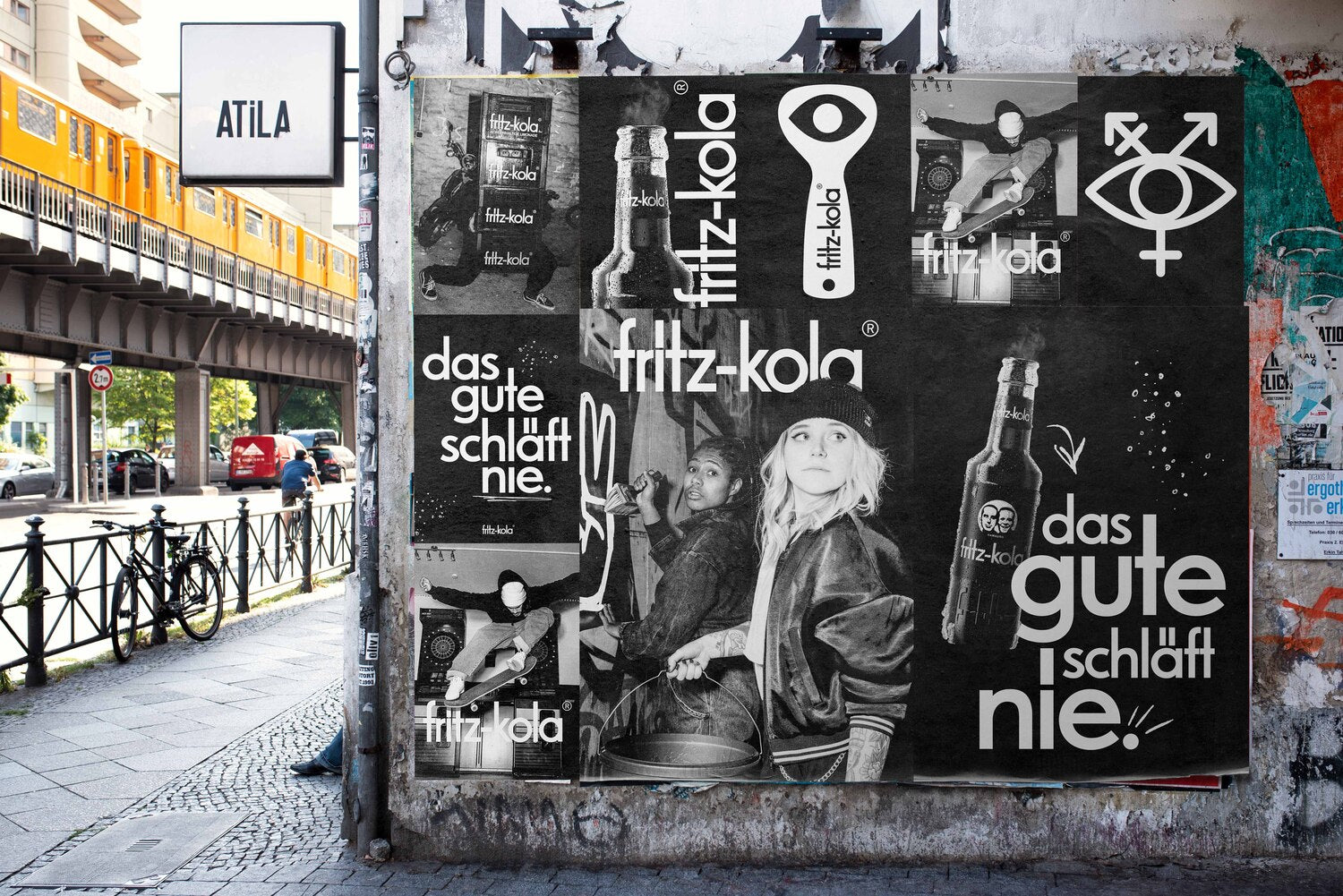 große schwarz-weiße plakatwand mit fritz-kola motiven und schriftzügen an einer hauswand, straße mit geländer und gelber hochbahn im hintergrund