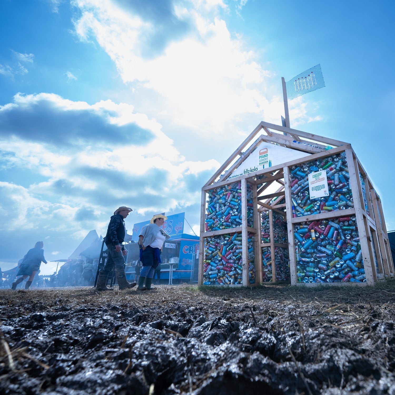 holzkonstruktion in form eines hauses gefüllt mit leeren dosen und flaschen steht auf einem festivalgelände, zwei personen gehen daran vorbei, himmel mit wolken im hintergrund  Geben Sie Ihr Feedback zu BizChat ab.