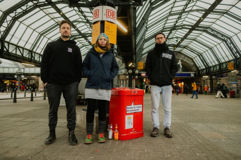 drei personen stehen auf einem bahnsteig neben einem roten mülleimer, mehrere glasflaschen stehen am boden davor, bahnhofshalle und menschen im hintergrund