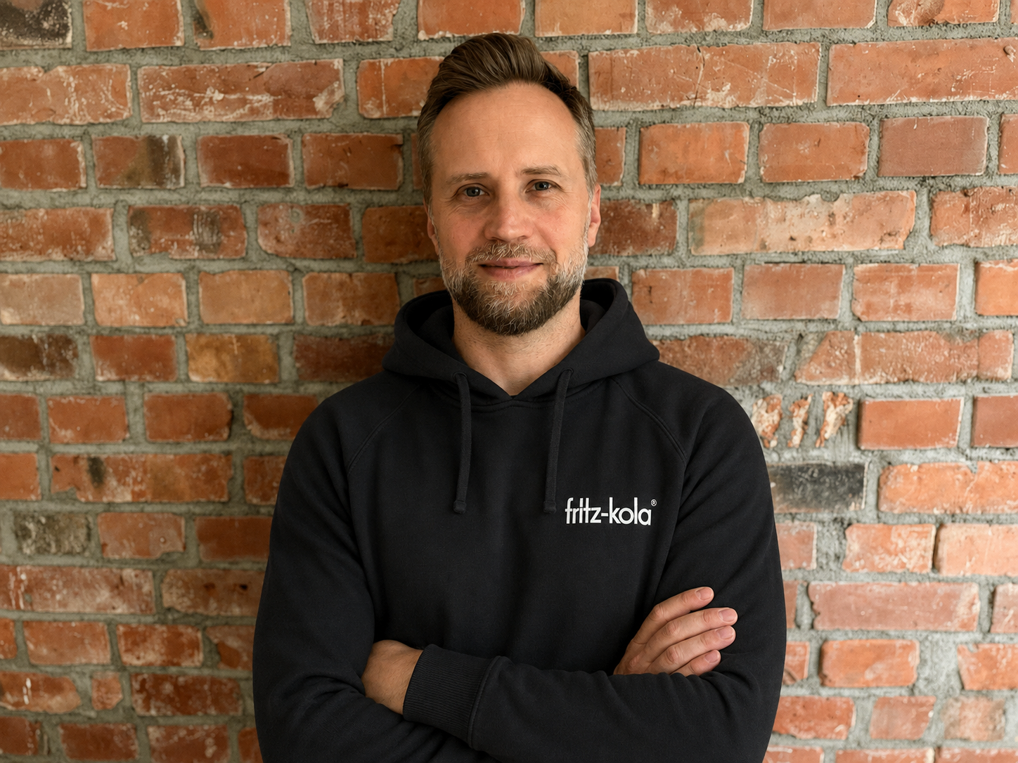 A portrait of Marcin Piaseczny, male person, in a fritz-kola hoodie in front of a brick wall.