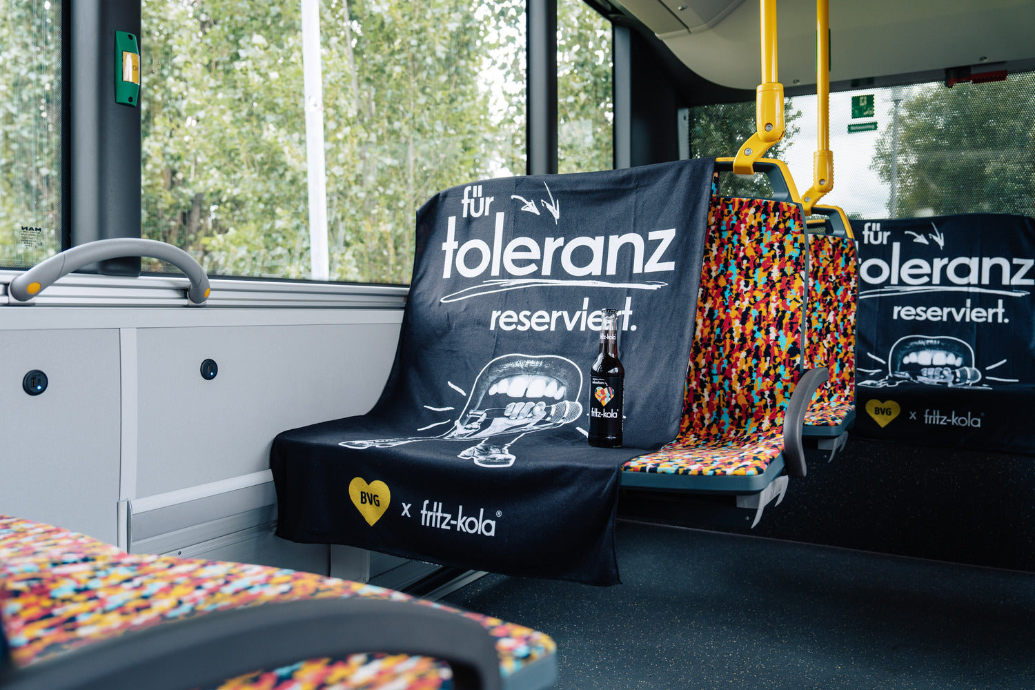 sitzplatz in einer straßenbahn ist mit schwarzem überwurf mit schriftzug „für toleranz reserviert“ versehen, fritz-kola glasflasche steht auf der sitzfläche, bunte sitzpolster und fenster im hintergrund