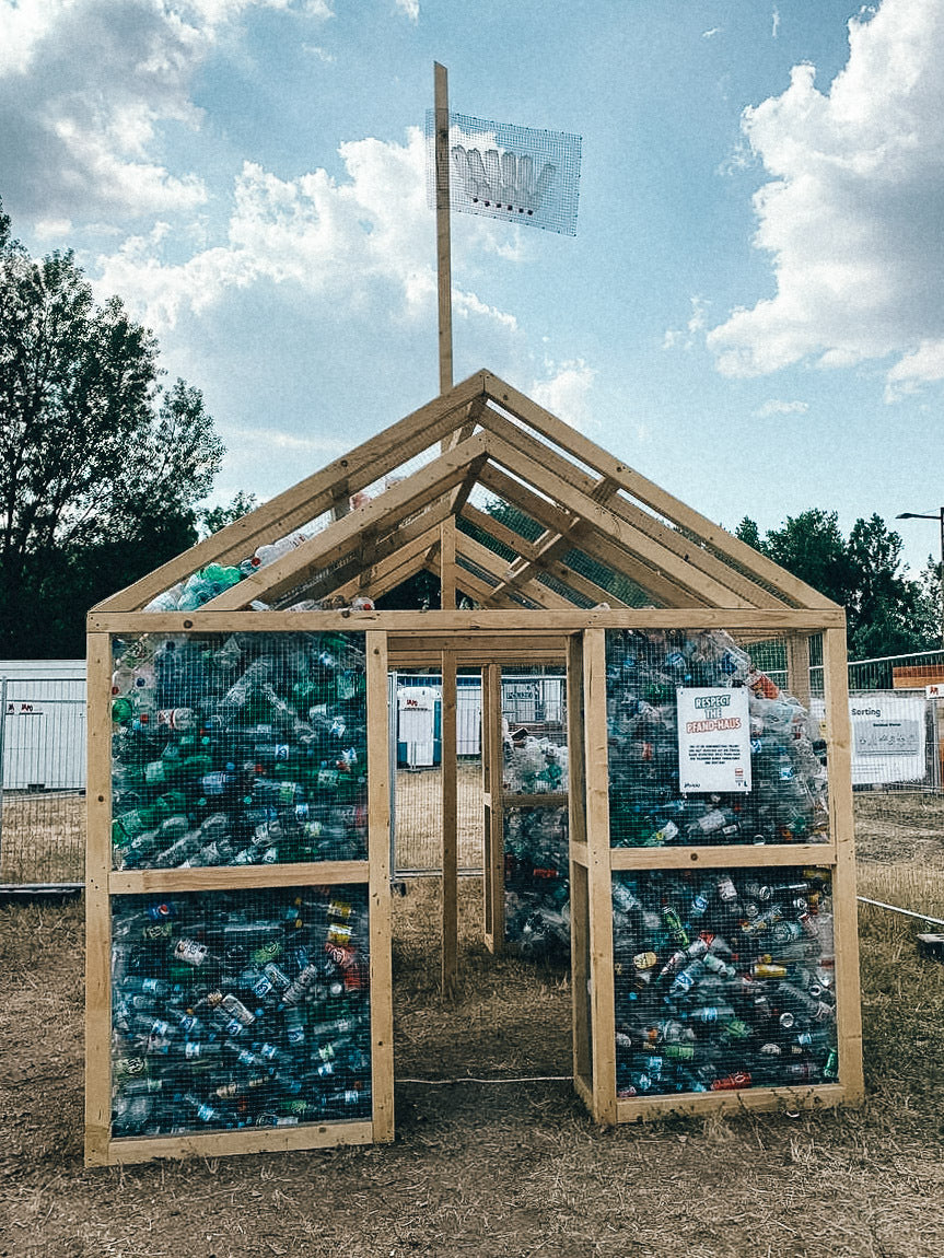 kleine holzkonstruktion in hausform mit drahtgitterwänden gefüllt mit leeren flaschen und dosen steht auf einem offenen gelände, fahne auf dem dach, himmel mit wolken und bäume im hintergrund