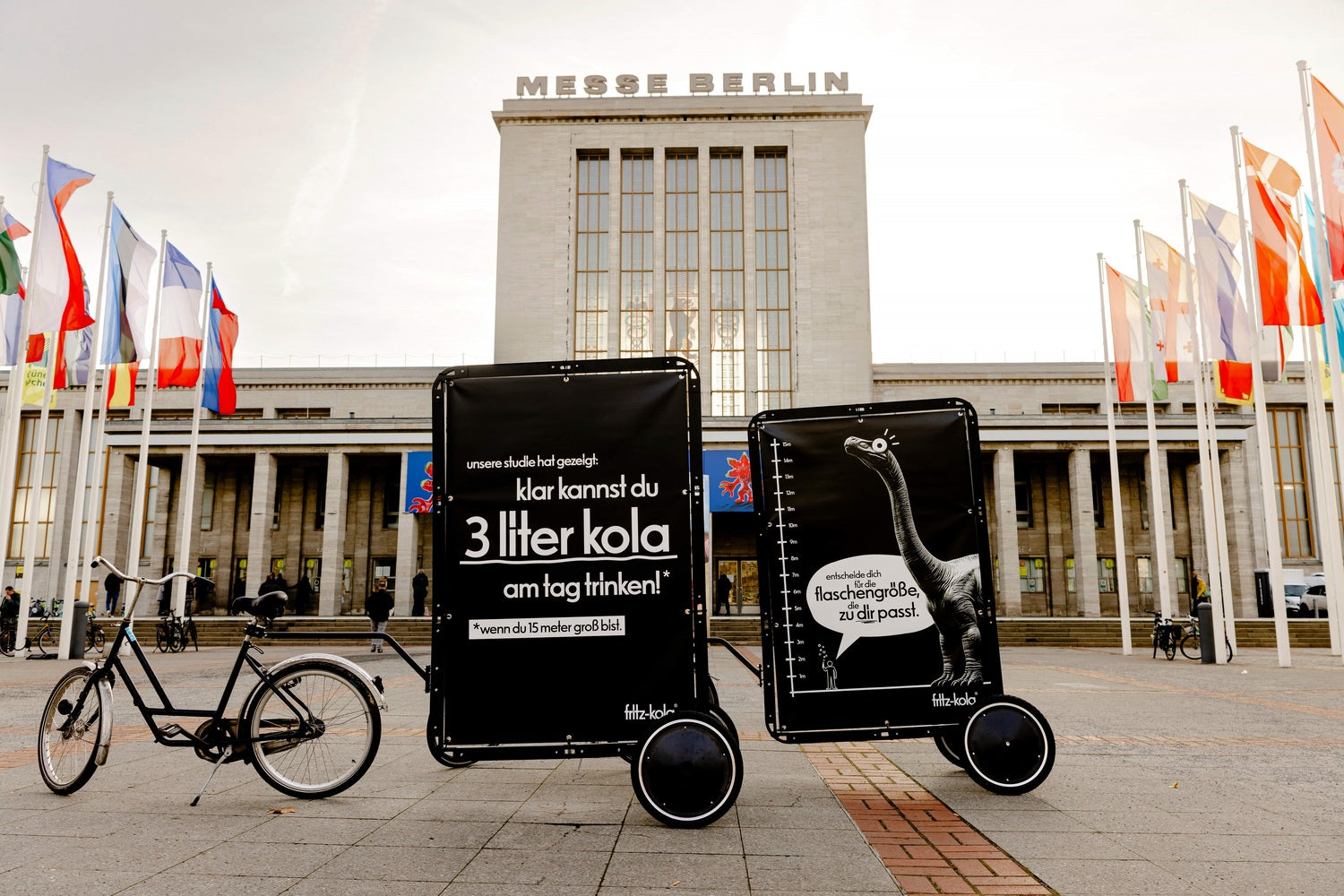 fahrrad mit zwei schwarzen anhängern mit fritz-kola plakaten steht vor einem gebäude mit der aufschrift „messe berlin“, flaggen links und rechts, pflasterplatz im vordergrund