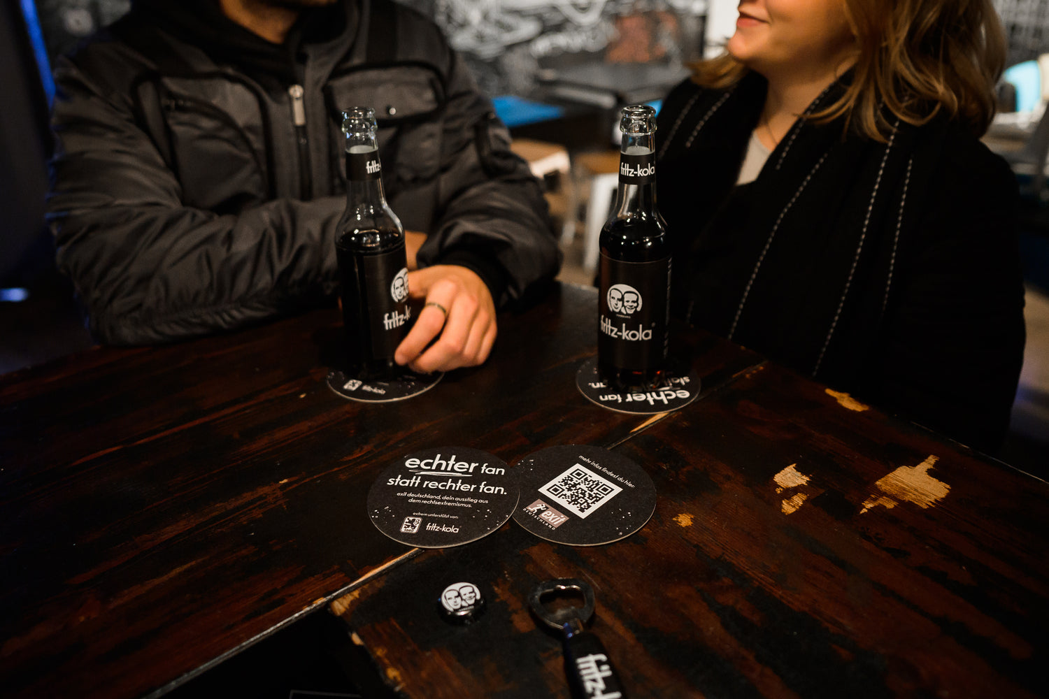 zwei personen sitzen an einem holztisch in einer bar, vor ihnen stehen zwei fritz-kola glasflaschen auf untersetzern, bierdeckel mit schrift und qr-code sowie flaschenöffner liegen auf dem tisch