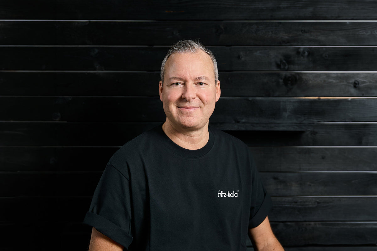 David Rodiek mit kurzen grauen Haaren im schwarzen fritz-kola T-Shirt vor schwarzer Wand.