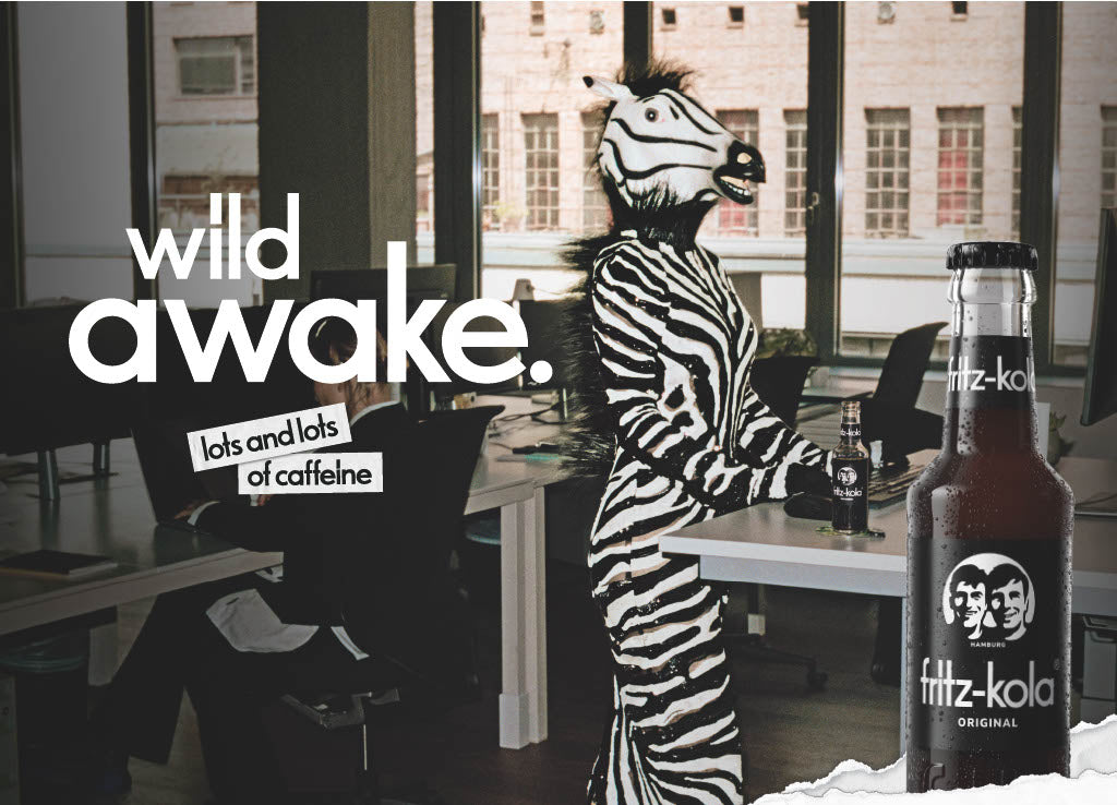 fritz-kola campagnebeeld met een persoon in zebrapak op kantoor, met de slogan „wild awake. lots and lots of caffeine“ en een fles fritz-kola original op de voorgrond.