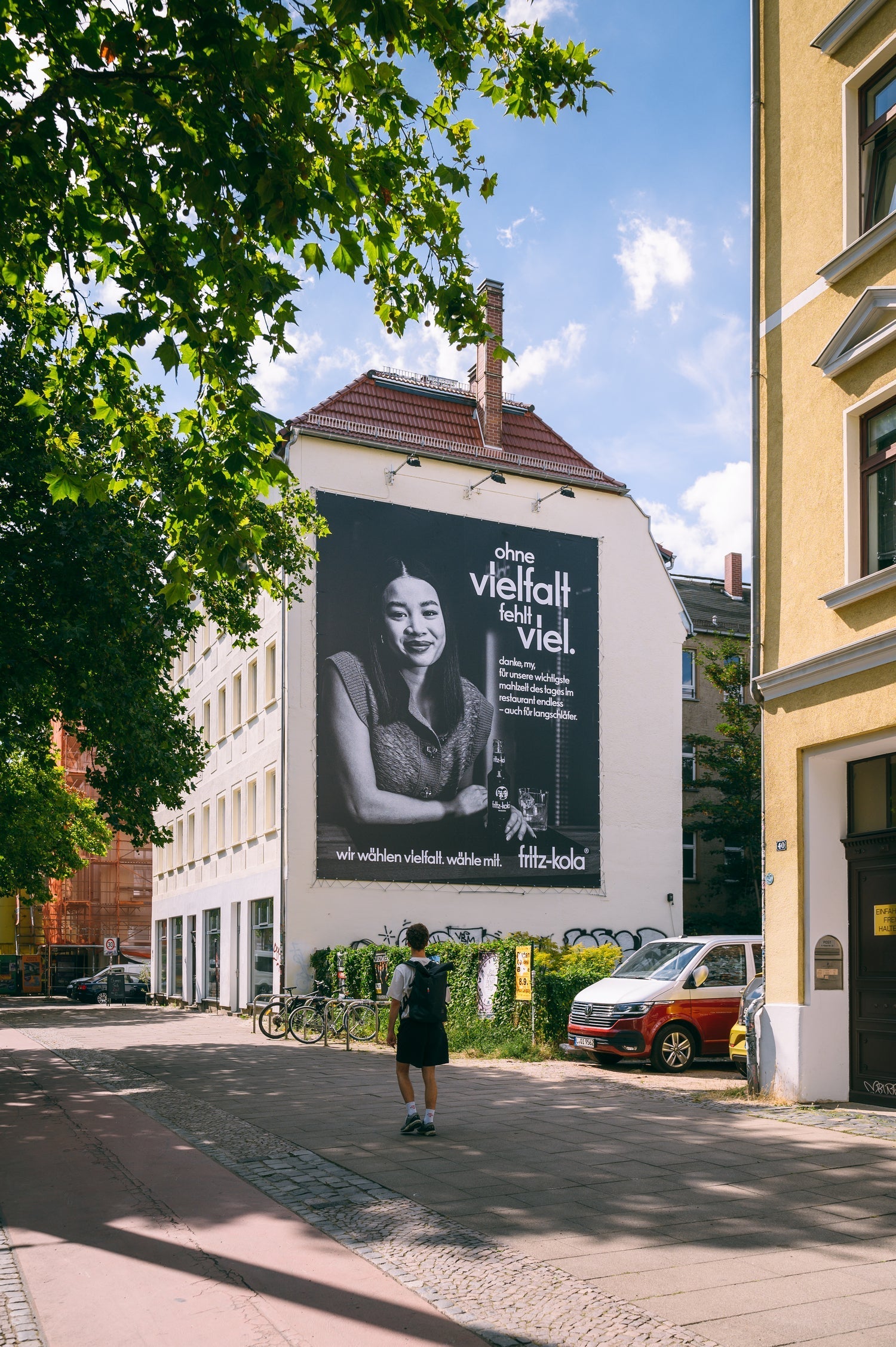 gebäude an einer straßenecke mit großem schwarz-weiß plakat an der fassade, person mit rucksack geht auf dem gehweg davor, bäume, fahrräder und parkende autos entlang der straße