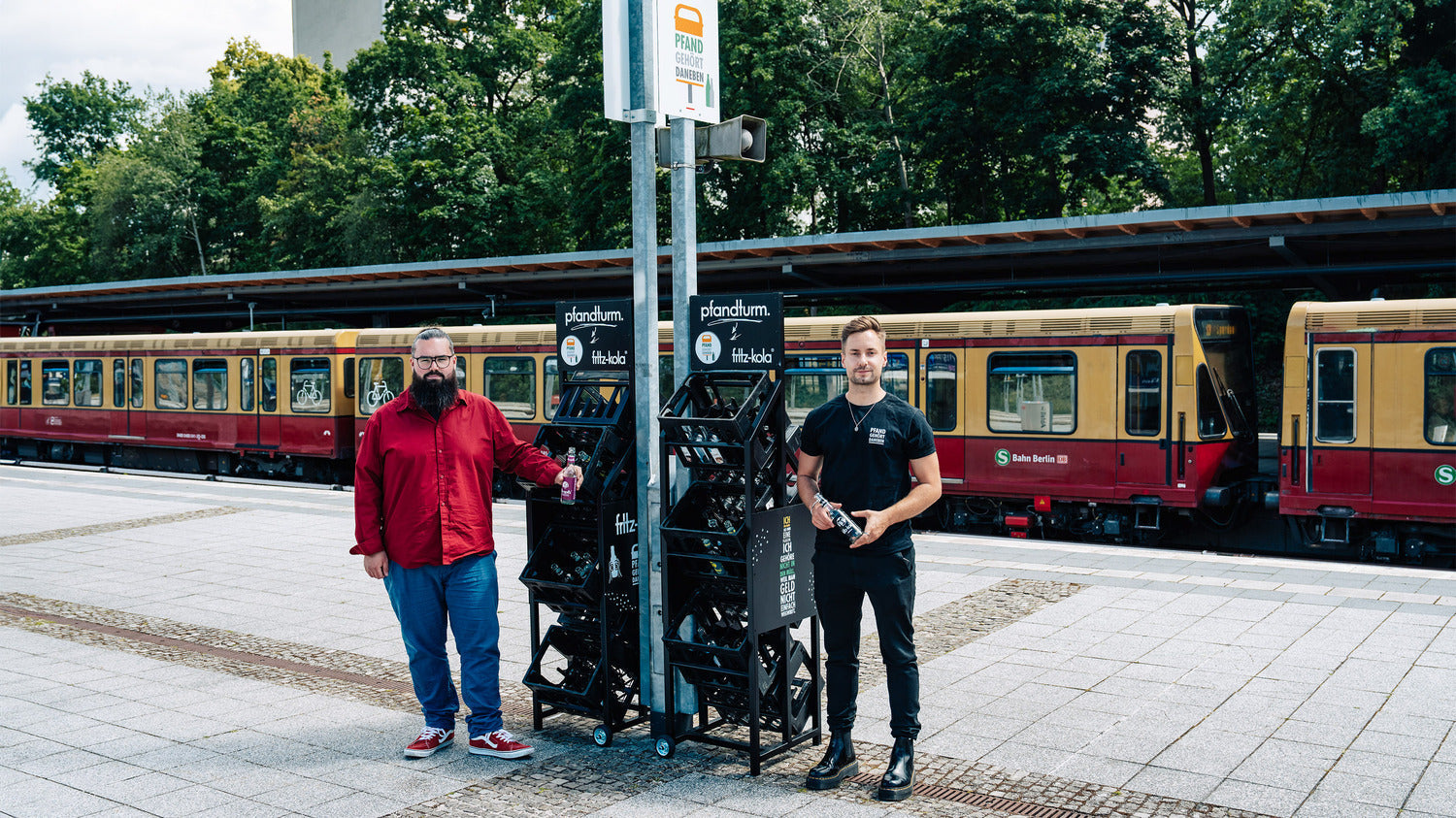 zwei personen stehen an einem bahnsteig neben einem pfandturm mit mehreren behältern für flaschen, s-bahn im hintergrund und bäume hinter dem gleis