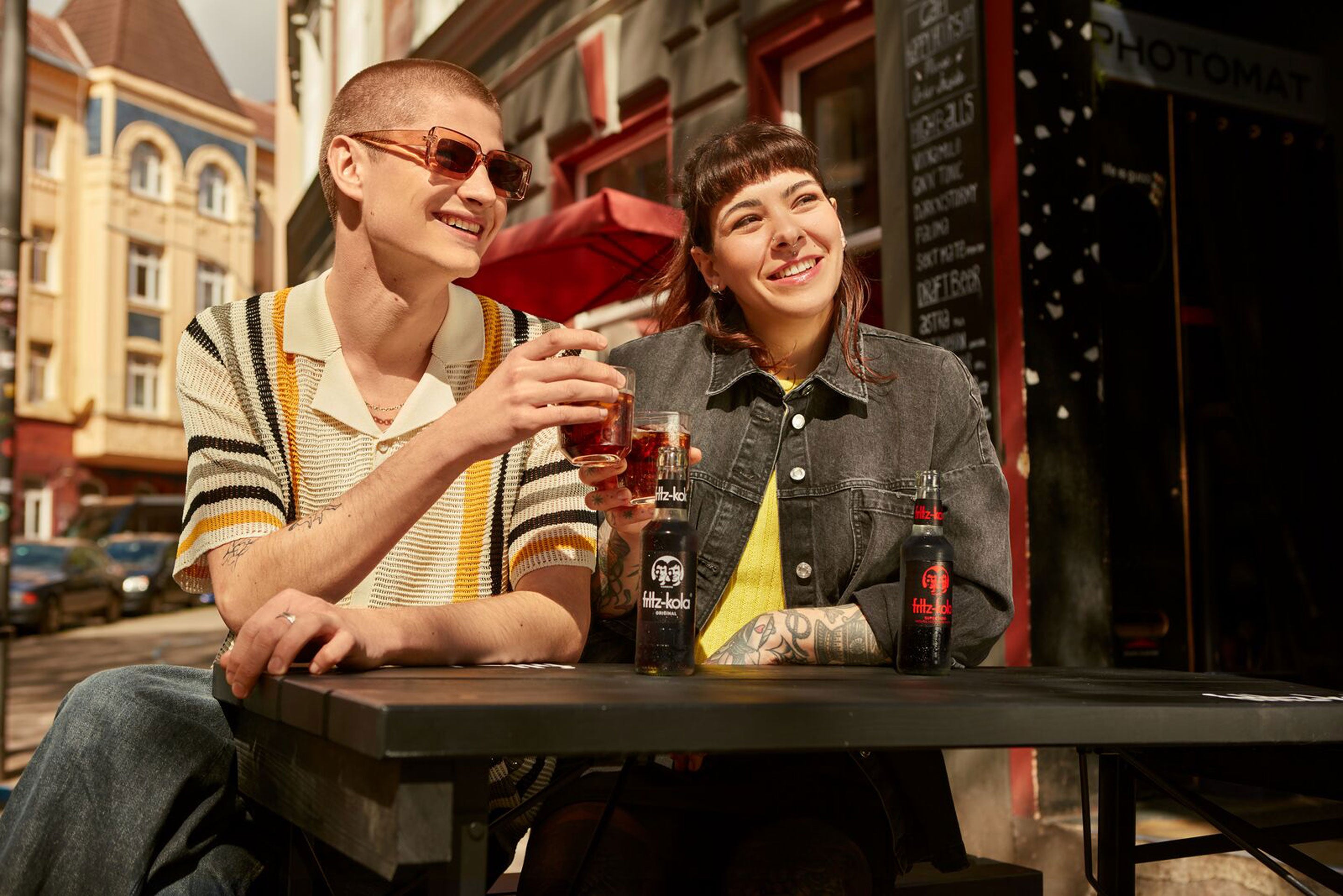Zwei lächelnde Freunde sitzen an Café-Tisch mit fritz-kola Flaschen. Links der Mann hat kurze Haare, Sonnenbrille und gestreiften Pullover. Rechte Frau trägt graue Jeansjacke über gelbem Top, hat Tattoos am Arm. Warmes Sonnenlicht, Straßencafé-Atmosphäre. sie stoßen an mit fritz-kola