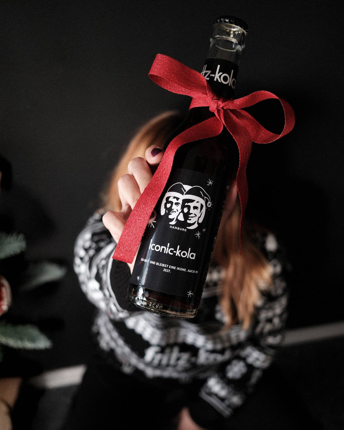 Eine Person hält eine dunkle fritz-kola Glasflasche vor die Kamera. Um den Flaschenhals ist eine große rote Schleife gebunden. Auf dem schwarzen Etikett sind das fritz-kola Logo, zwei illustrierte Gesichter mit Weihnachtsmützen, kleine Sterne sowie der Schriftzug „iconic-kola“ und „Du bist und bleibst eine Ikone. Auch in 2027.“ zu sehen. Die Person und der dunkle Hintergrund sind unscharf, sodass die Flasche mit der roten Schleife im Mittelpunkt steht.