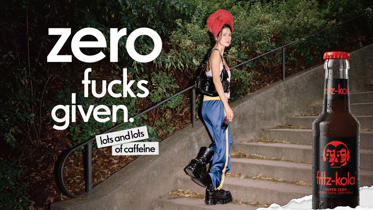 Person with a red towel wrapped around the head walking up outdoor stairs with a bottle of fritz-kola Super Zero. Campaign visual featuring the slogan “zero fucks given” and a large product bottle.