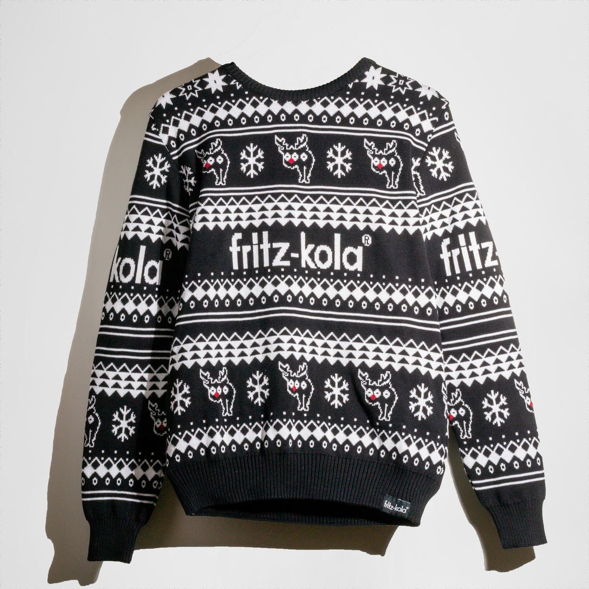 Ein weihnachtlicher fritz-kola Strickpullover im klassischen 'Ugly Christmas Sweater' Design, fotografiert vor weißem Hintergrund. Der schwarze Pullover zeigt ein skandinavisches Strickmuster mit weißen Rentieren, Schneeflocken und dem fritz-kola Logo. 