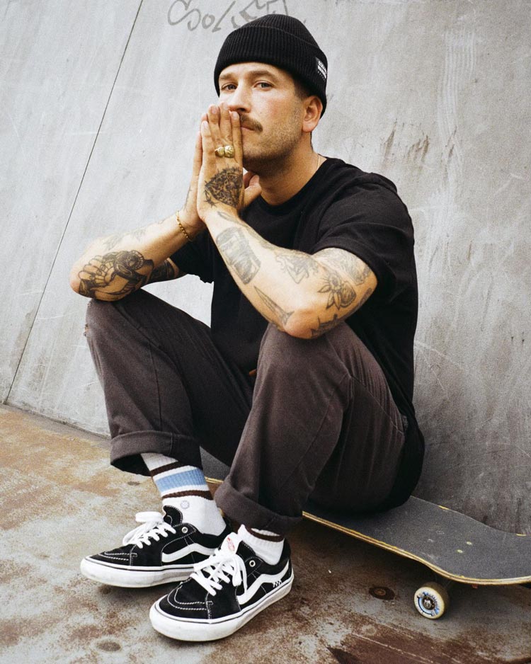 Lifestyle Aufnahme des Künstlers BRODERSON. Er sitzt auf einem Skateboard, trägt ein schwarzes Shirt und Beanie sowie Sneaker. An den armen trägt er Tattoos. 