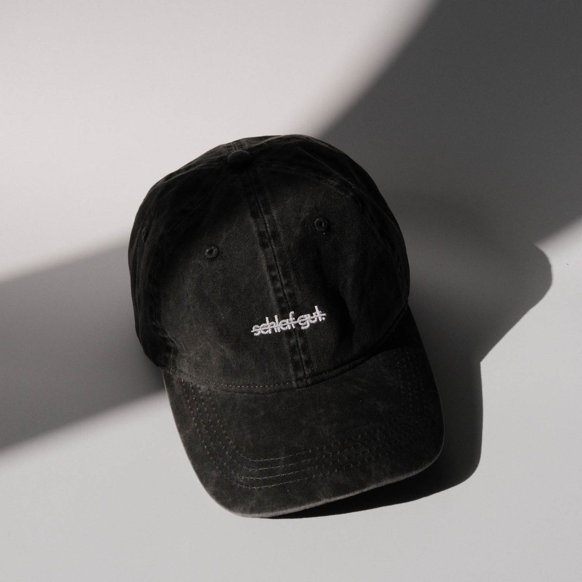 Schwarze Cap in verwaschener Optik mit weiß gesticktem und durchgestrichenem Schriftzug „schlaf gut.“ auf der Vorderseite, fotografiert vor hellem Hintergrund mit weichen Licht- und Schattenflächen.