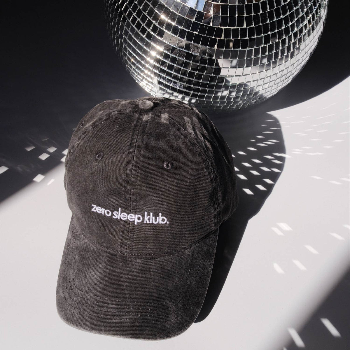 Schwarze Cap mit weiß gesticktem Schriftzug „zero sleep klub.“ vor einer silbernen Discokugel. Lichtreflexe der Discokugel fallen auf Cap und Hintergrund und erzeugen eine Club-Atmosphäre.