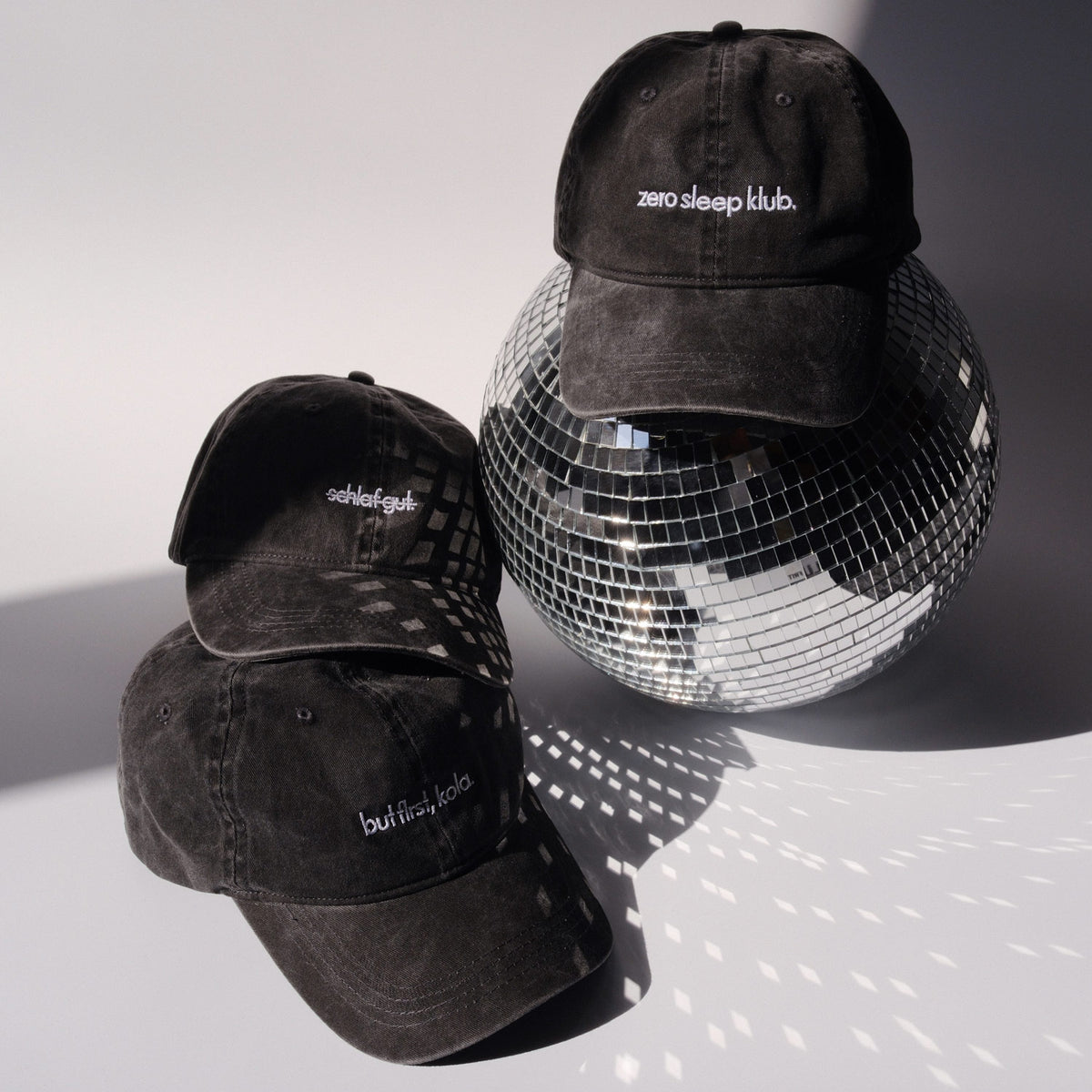 Drei schwarze Caps mit weißer Stickerei „but first, kola“, „schlaf gut.“(durchgstrichen) und „zero sleep klub.“ arrangiert neben einer silbernen Discokugel auf hellem Hintergrund.