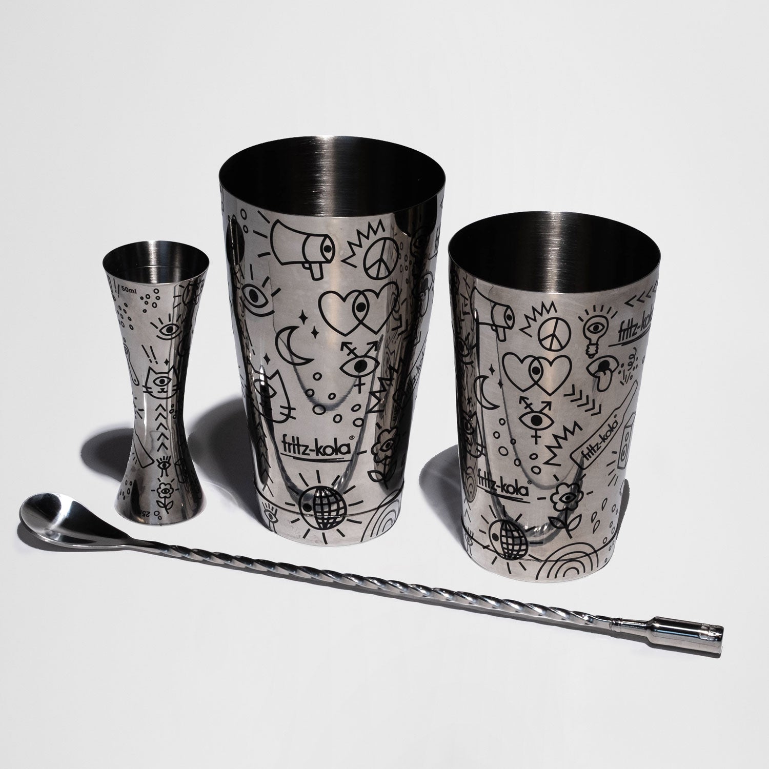 cocktailset mit zwei metallbechern, einem jigger und einem langen rührlöffel mit fritz-kola muster vor weißem hintergrund