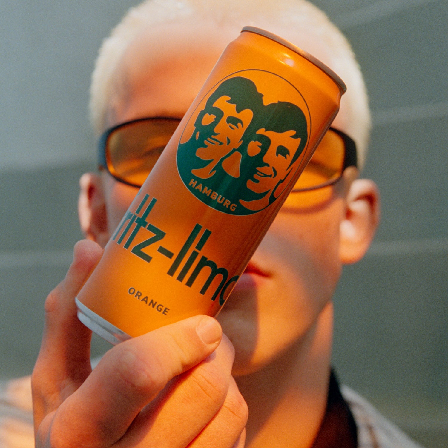 Person hält eine Dose fritz-limo Orange vor das Gesicht. Die orangefarbene Dose steht im Fokus, während die Person mit orange getönten Sonnenbrillen im Hintergrund unscharf zu sehen ist.